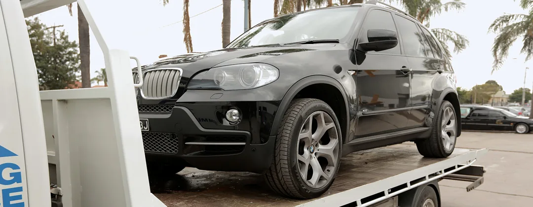 A damaged BMW X5 on a tow truck being unloaded outside the workshop.